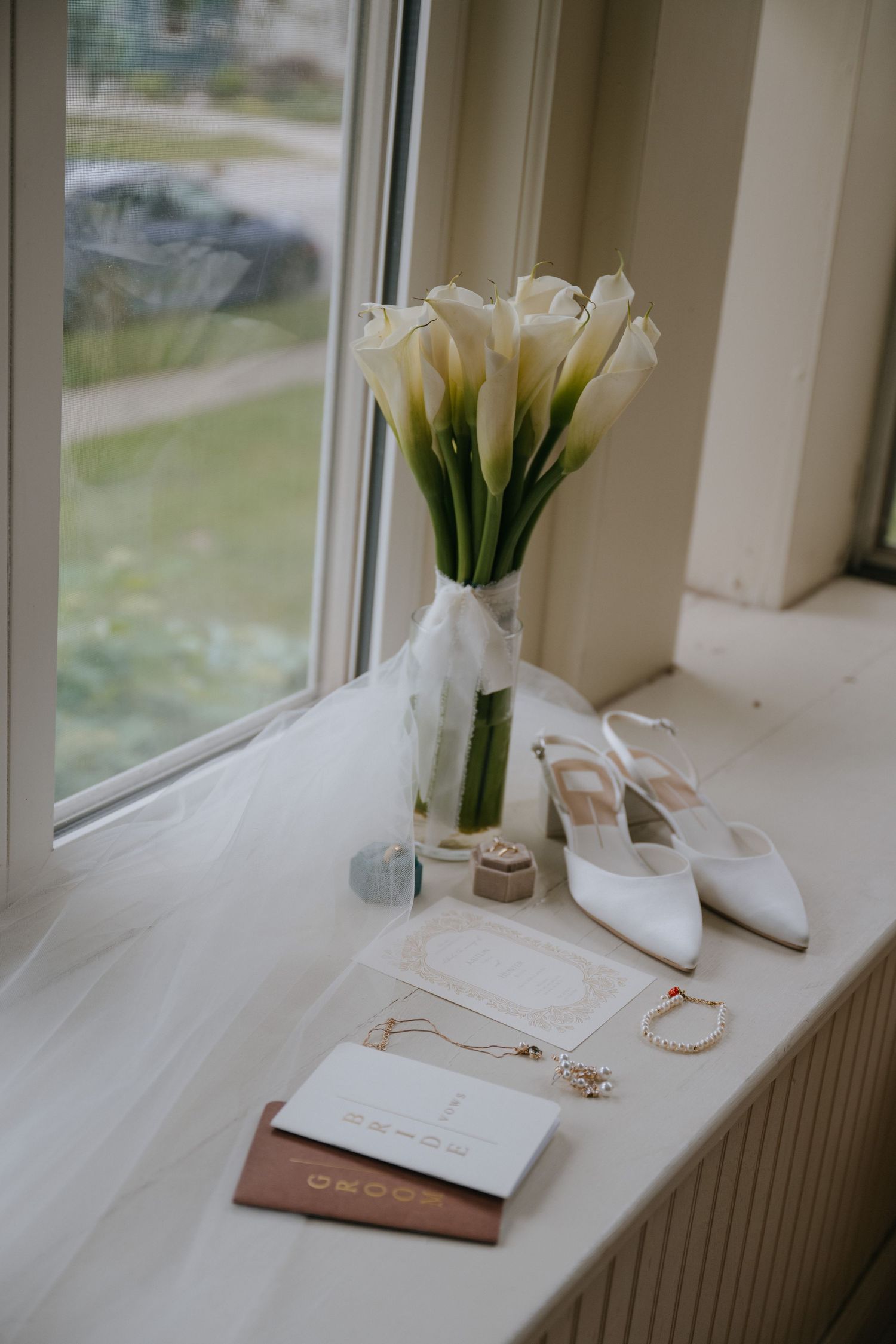Wedding details with calla lily bouquet, jewelry and stationery arranged on a sunny windowsill.