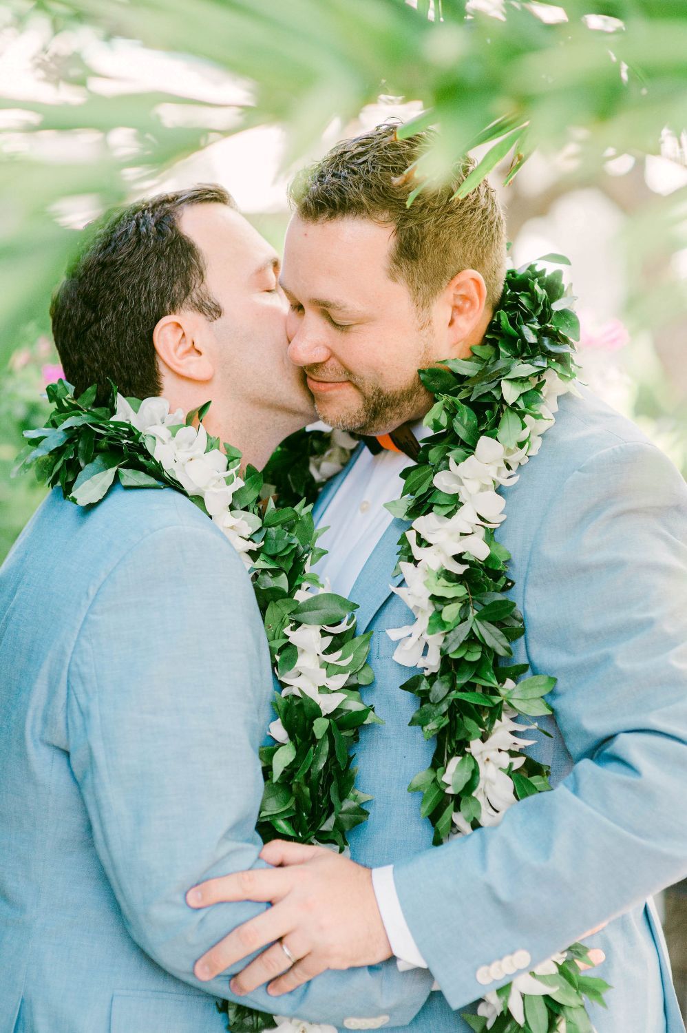 A romantic moment shared between two people wearing blue suits and floral leis in a garden setting.