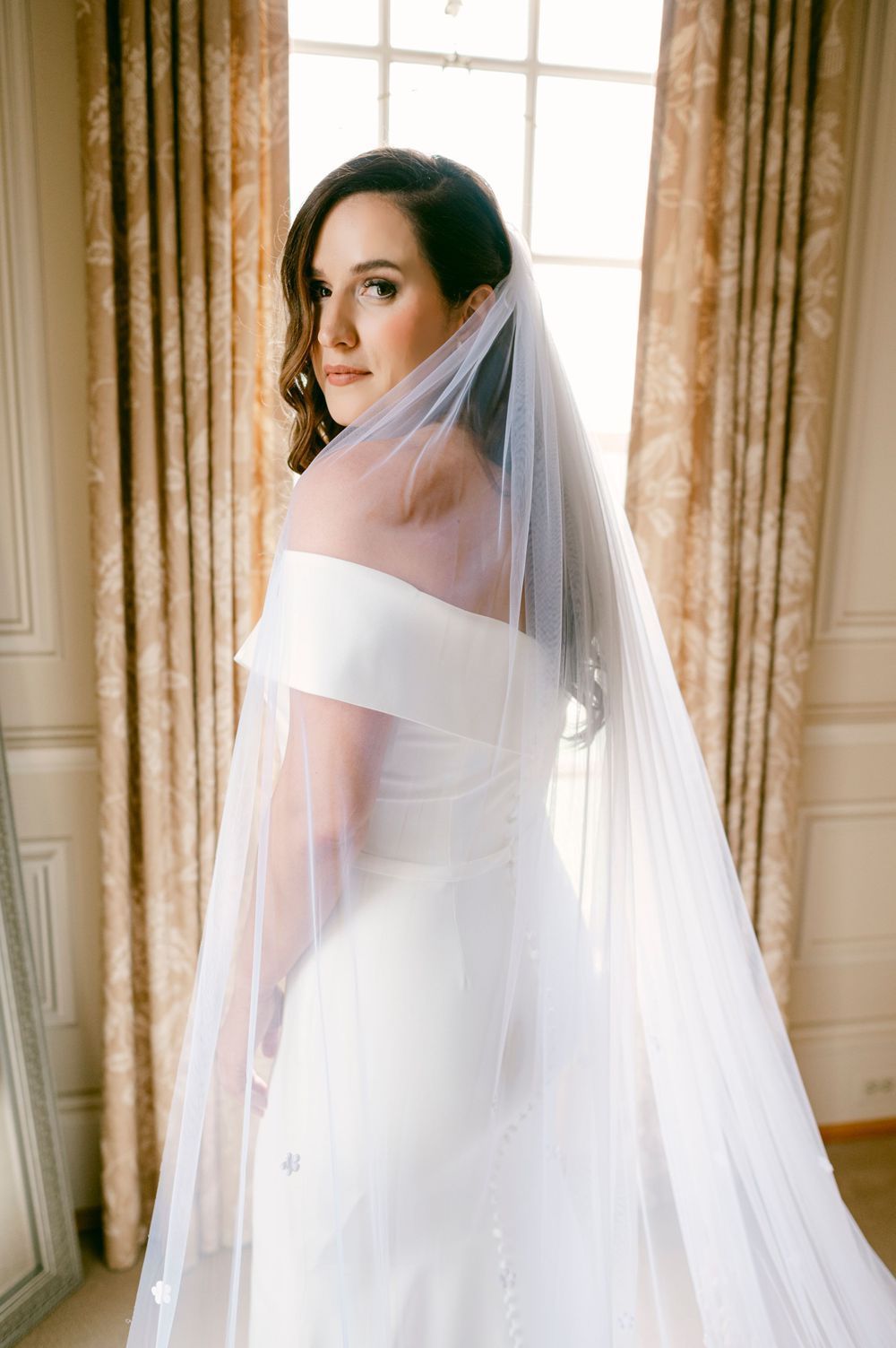 Elegant bride in off-shoulder white wedding gown and flowing veil poses by window with sheer curtains in natural light.