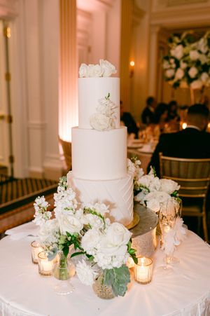Elegant white wedding cake with fresh floral decorations and glowing candlelight on white tablecloth at reception.