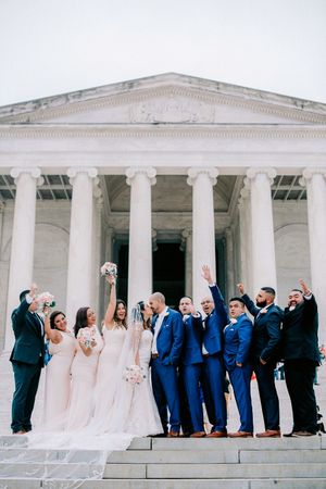 Wedding party in blush pink dresses and navy blue suits celebrate on steps of neoclassical building with white columns.