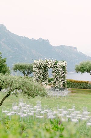 White chairs arranged on a green lawn for an outdoor wedding ceremony with blooming trees and mountains in the background.