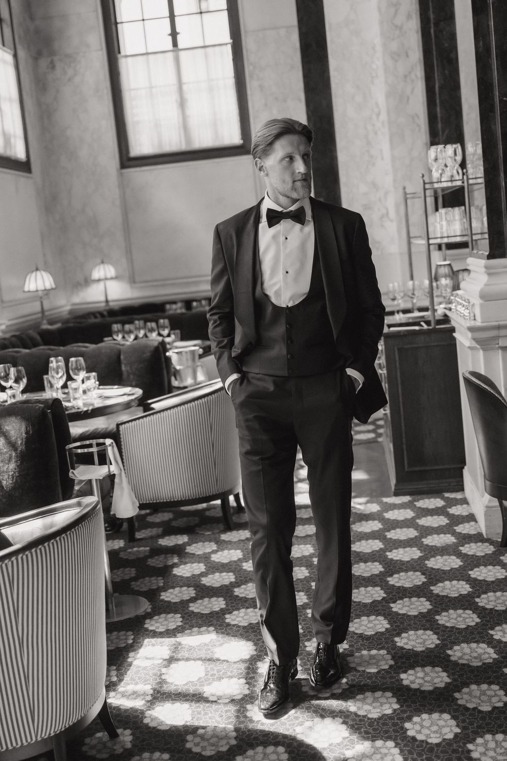 A man in a classic black tuxedo stands elegantly in a vintage restaurant setting with patterned floors and ambient lighting.
