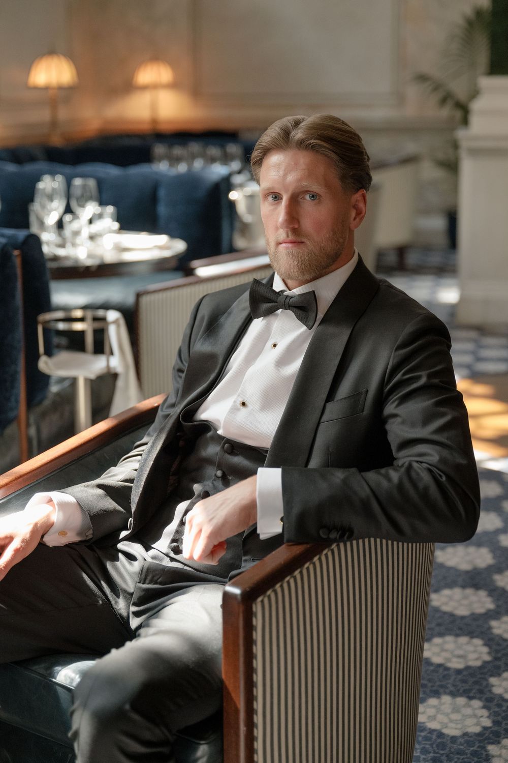 A well-dressed person in a black tuxedo and bow tie reclines in an elegant upholstered armchair.