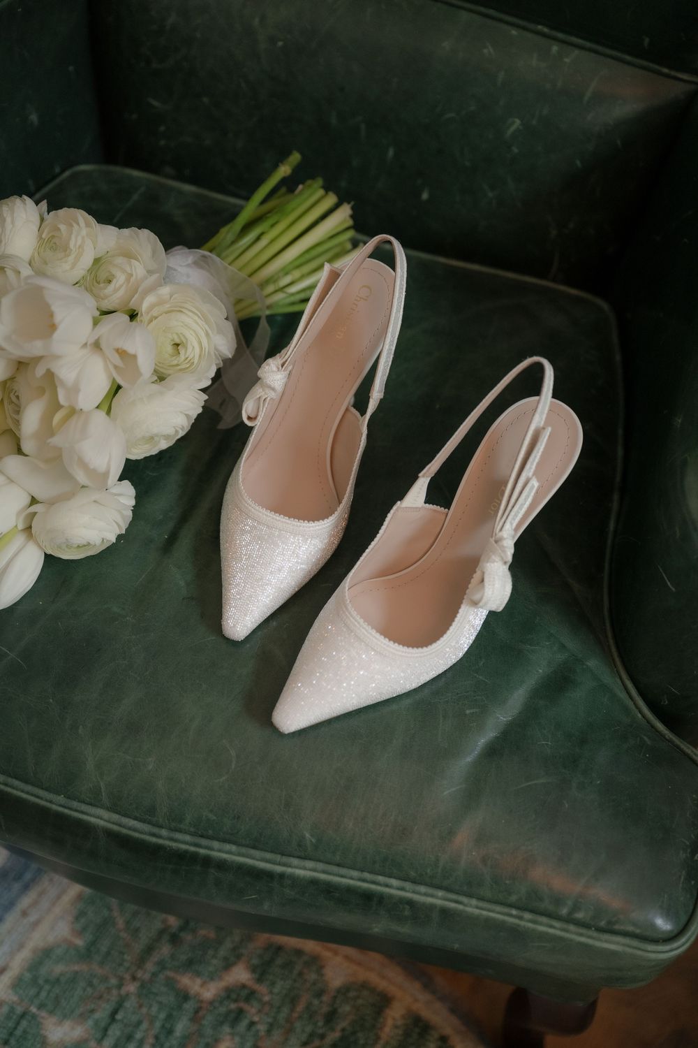 White bridal slingback heels with sparkly details rest beside a bouquet of white roses on a green velvet chair.