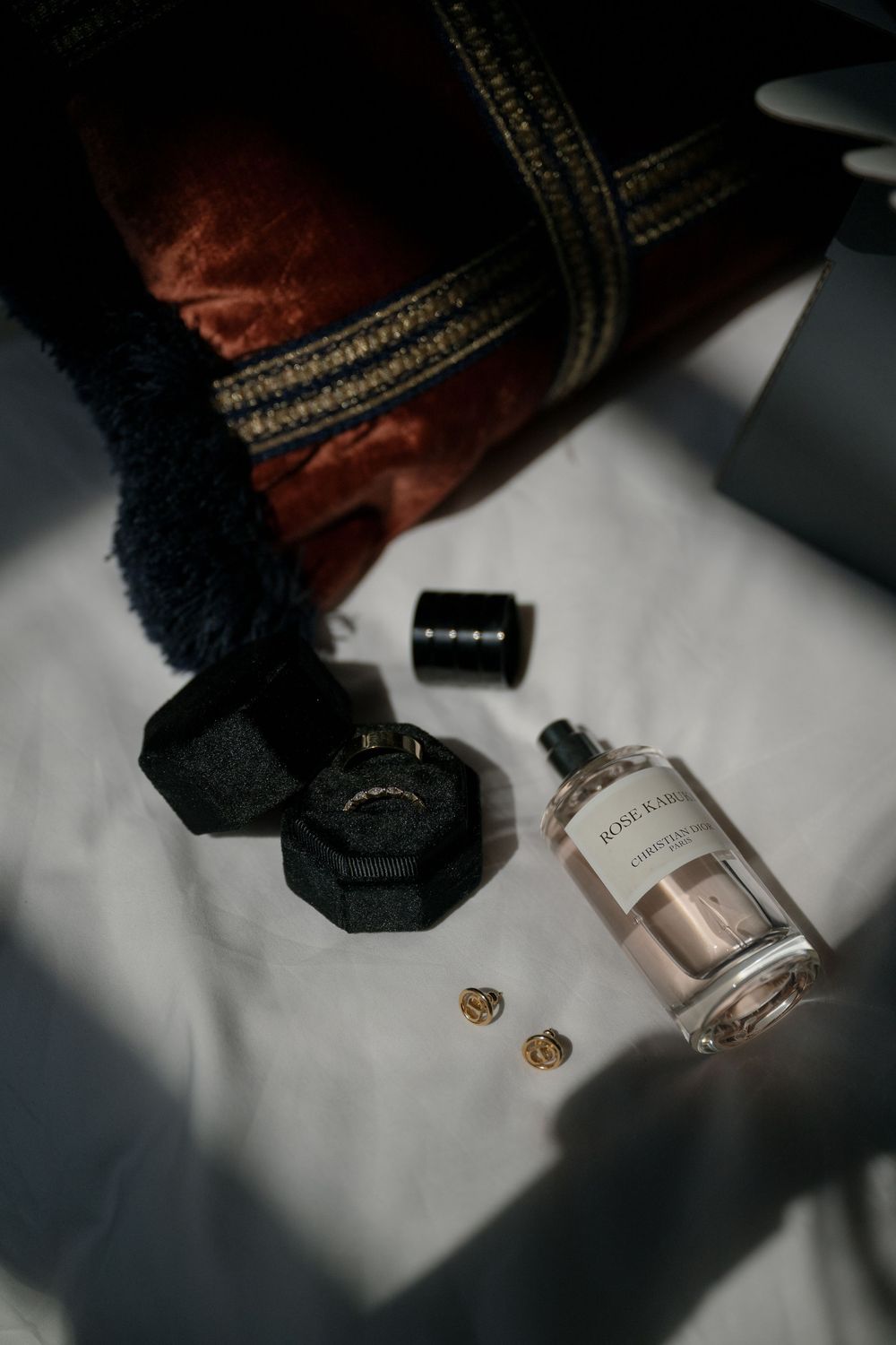Wedding accessories including rings, perfume bottle and velvet ring box arranged on white surface in dramatic lighting.