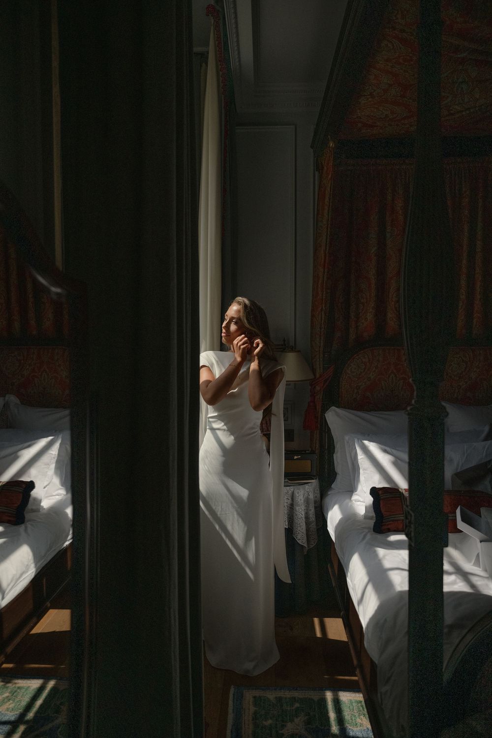 A bride in a white dress stands in dramatic window light between two beds in an elegant bedroom.