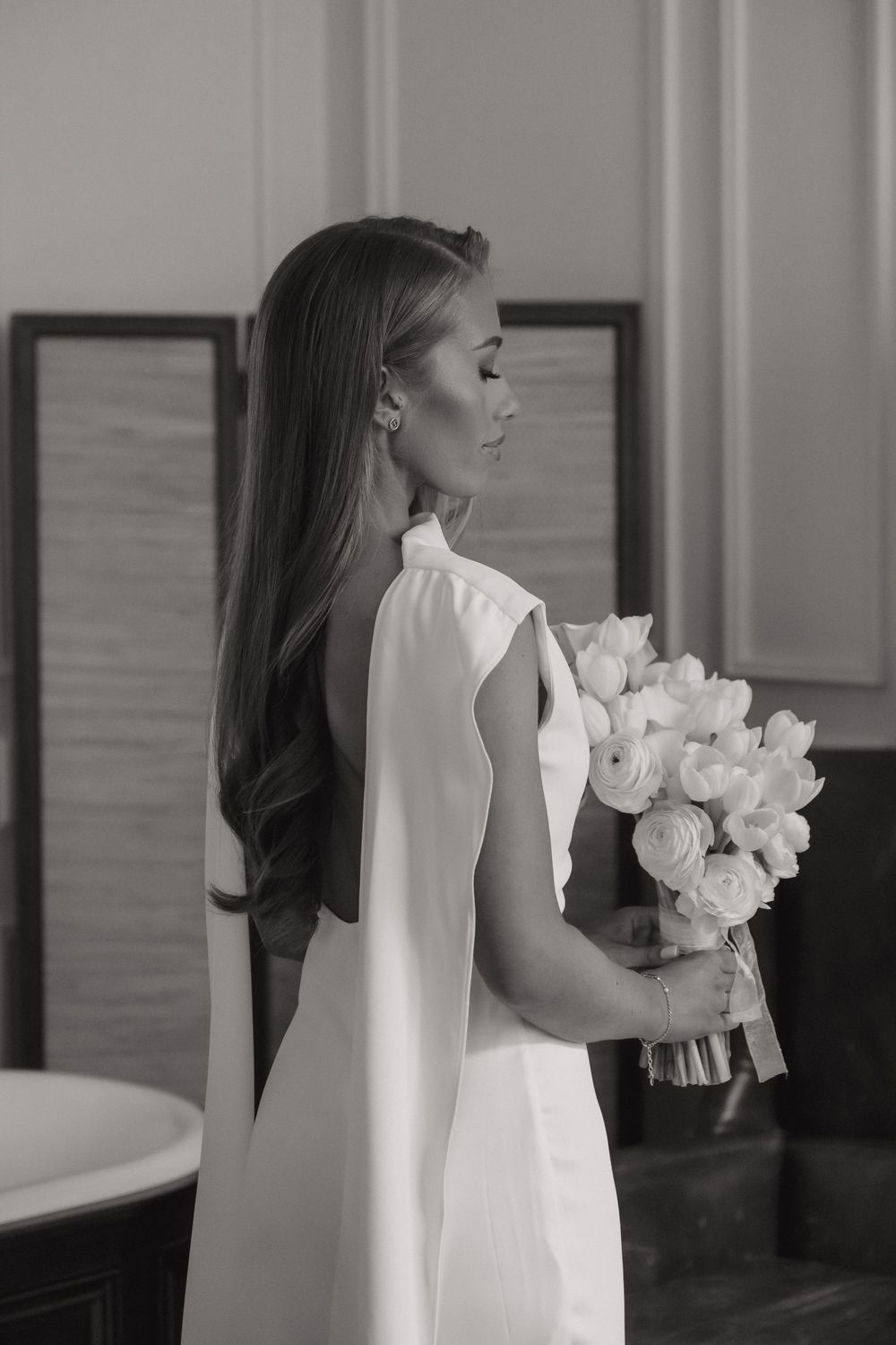 Elegant bride in a sleek white dress holds a bouquet of white roses against a light backdrop.