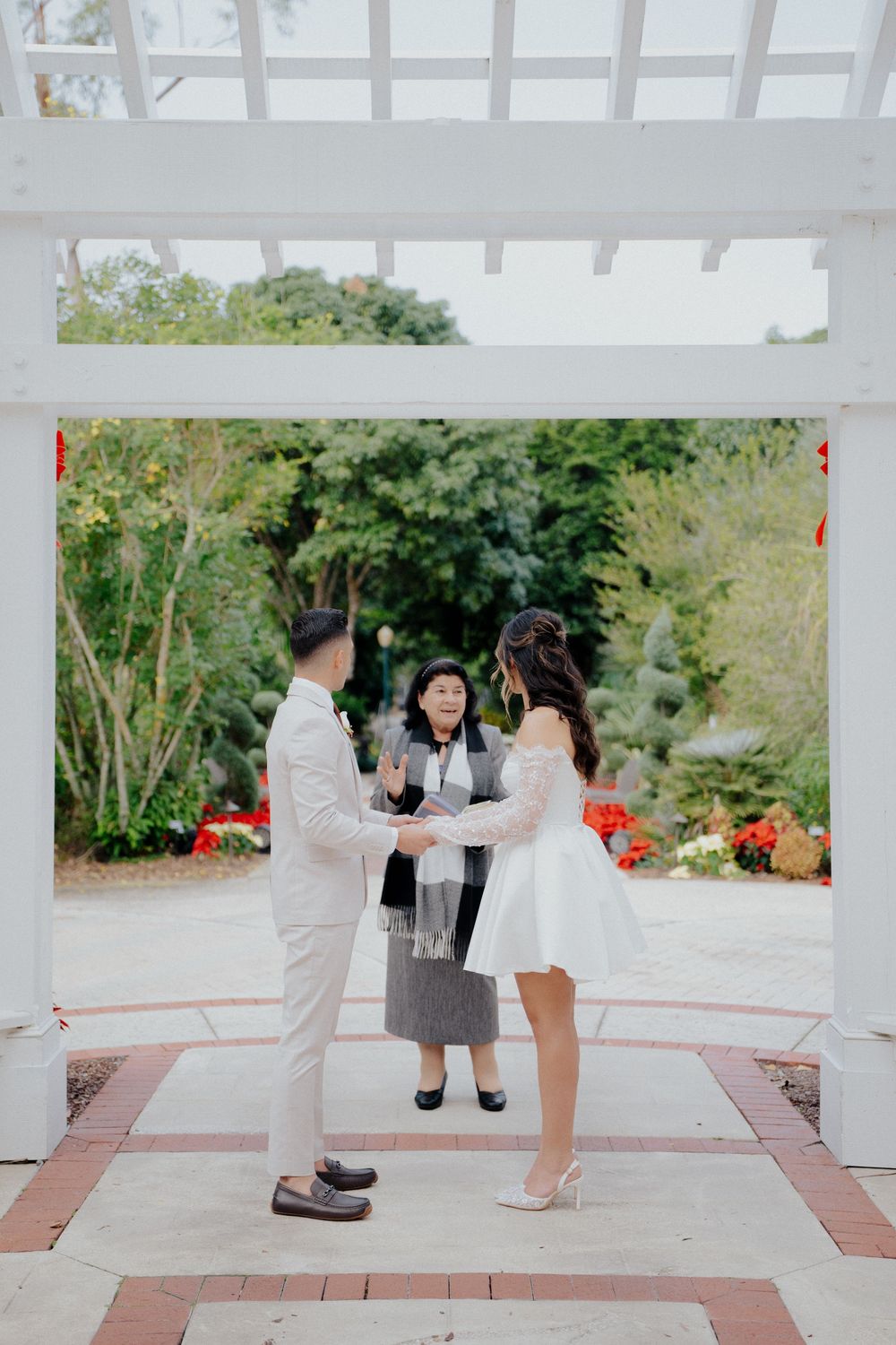 A couple shares an intimate moment on a white gazebo deck surrounded by lush greenery and red flowers.
