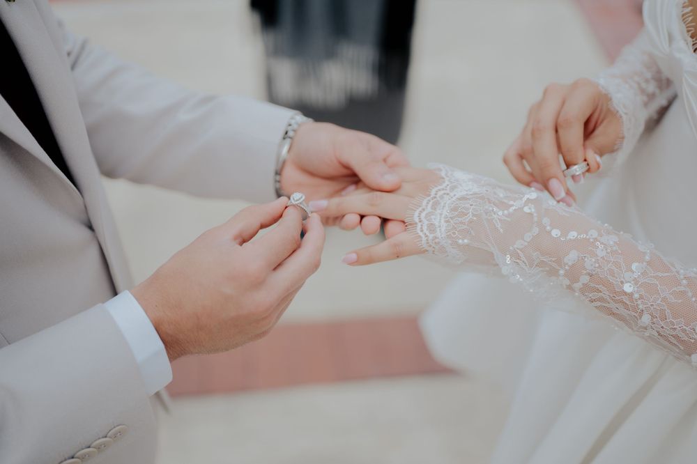 Close up of hands exchanging wedding rings during a ceremony with delicate lace sleeve detail.
