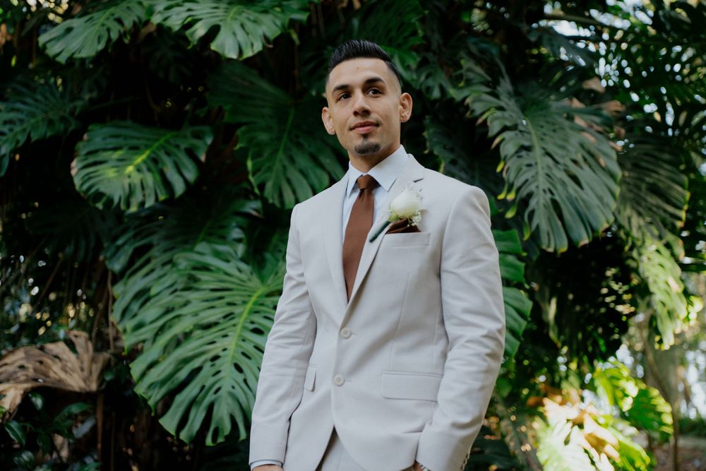 Groom in a crisp white suit jacket poses against tropical foliage background.
