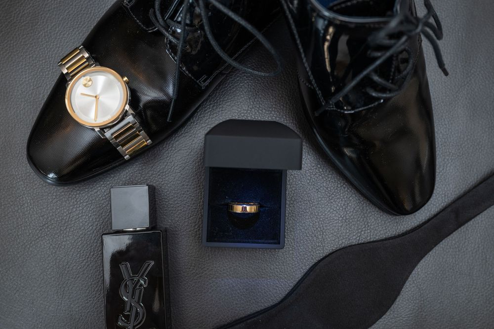 A groom's wedding accessories including black dress shoes, gold watch, cologne bottle, and engagement ring in navy box.