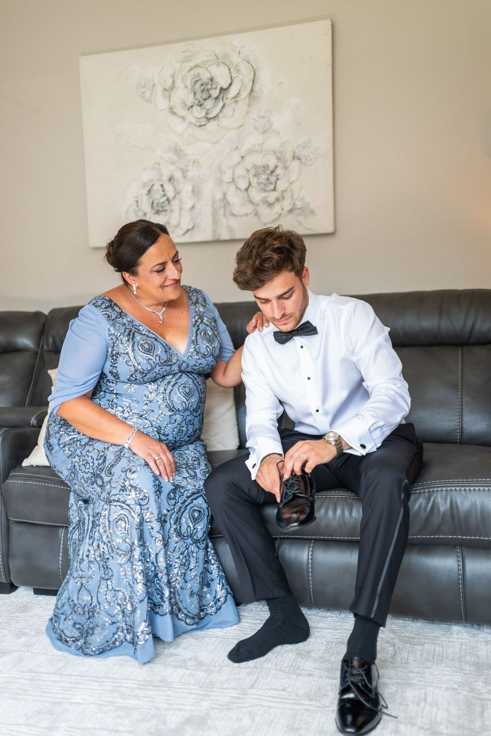 Two people in formal attire sit together on a leather couch beneath floral wall art in a living room setting.