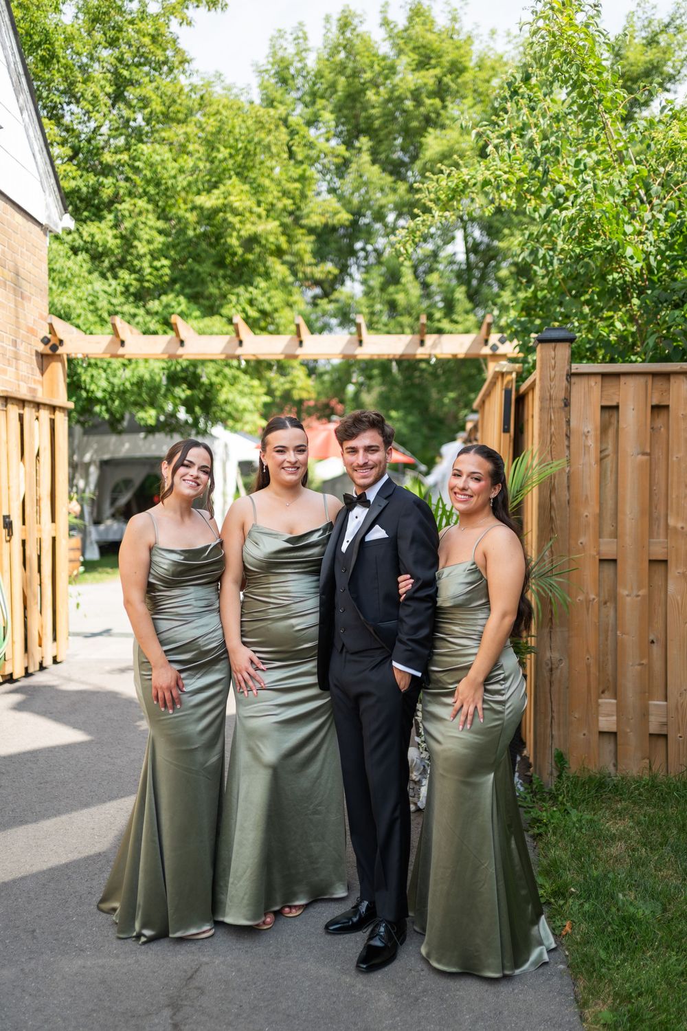 Wedding party members in sage green satin dresses and formal black tuxedo pose together in a backyard setting.