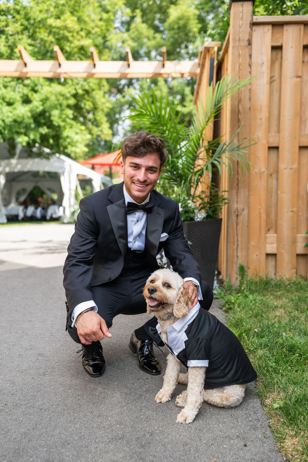 A person in a black tuxedo kneels down to pet a small white and brown dog on a paved path near a wooden fence.