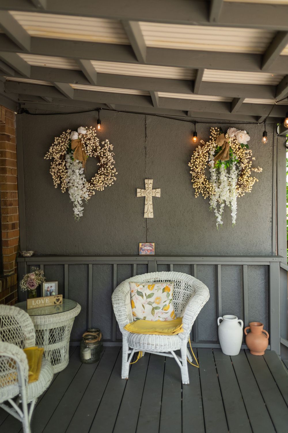A cozy porch corner with white wicker chair, decorative wreaths, and a cross mounted on dark gray paneled walls.