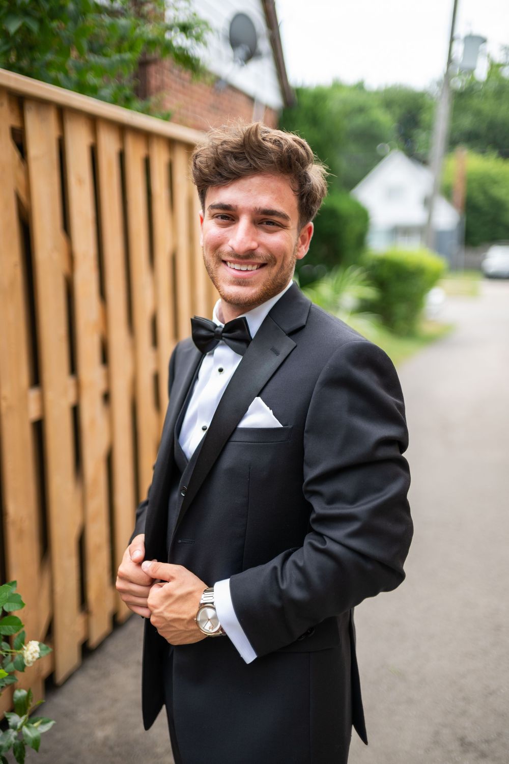 A groom in a classic black tuxedo smiles and poses outdoors against a wooden fence backdrop.