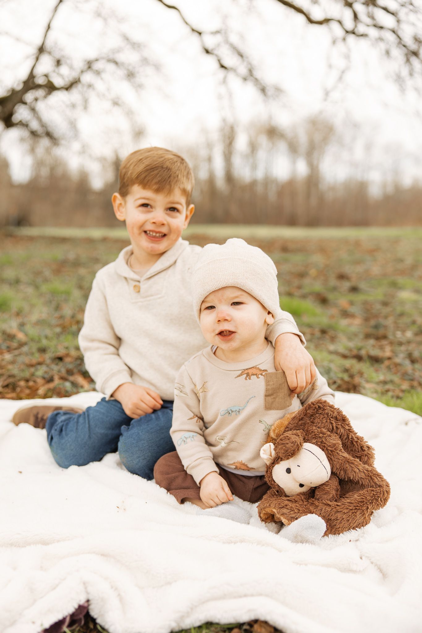 Two young brothers smile together on a white blanket outdoors, with a stuffed monkey toy nearby.