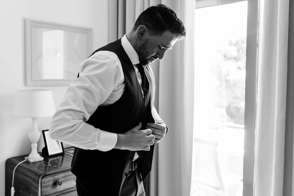 groom adjusting vest by window light during wedding preparations