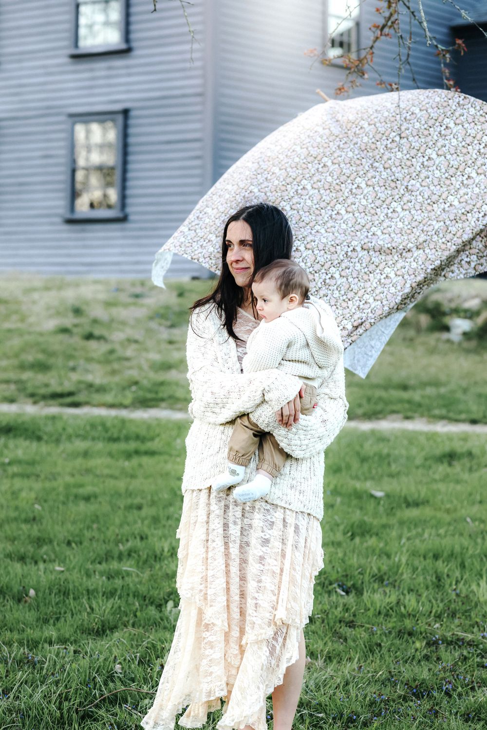 A mother with dark hair wearing a lace cream dress holds her baby boy wearing a cream sweater and tan pants in front of Nathan Meriam House