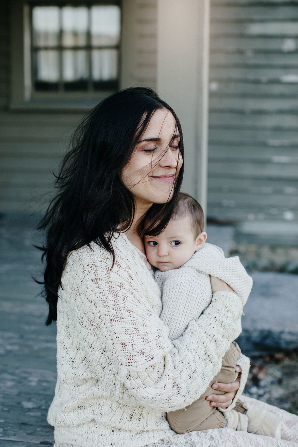 A mother wearing a cream sweater snuggles her baby boy close to her sitting on a weathered porch at the Buttricks House in Lincoln, MA