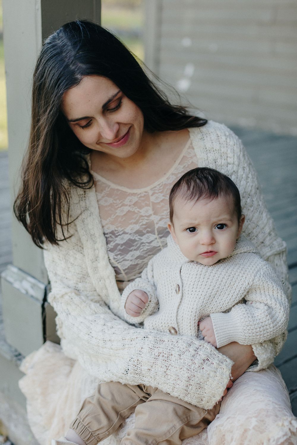A mother with brown hair wearing a cream sweater and lace dress holds her baby boy on a weathered porch in Lincoln, MA for motherhood photos