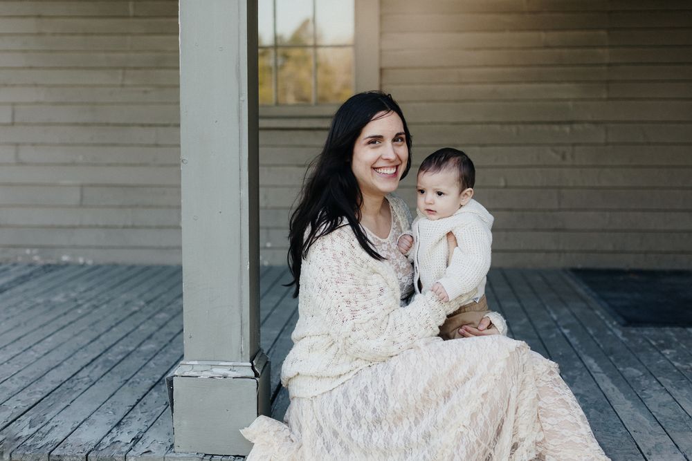 A mother wearing a cream sweater and lace dress holds her baby boy on her lap on a weathered porch at the Buttricks House in Lincoln MA