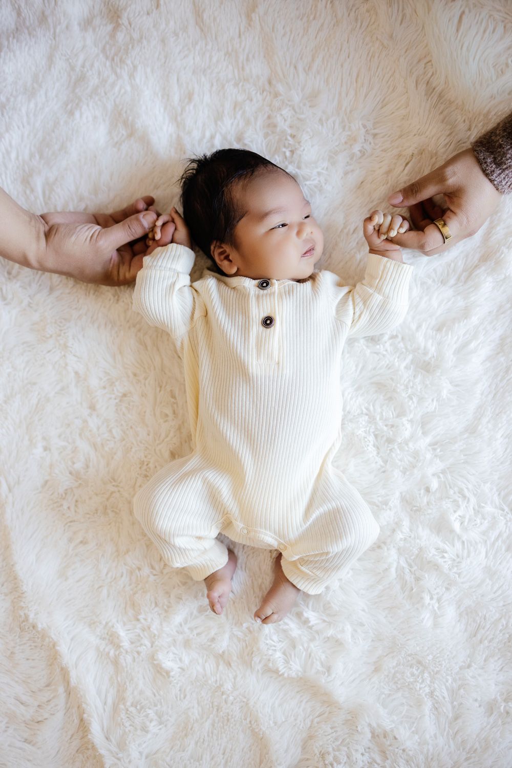 a month-old baby lays on a white blanket while holding his parents fingers at his new born photo shoot