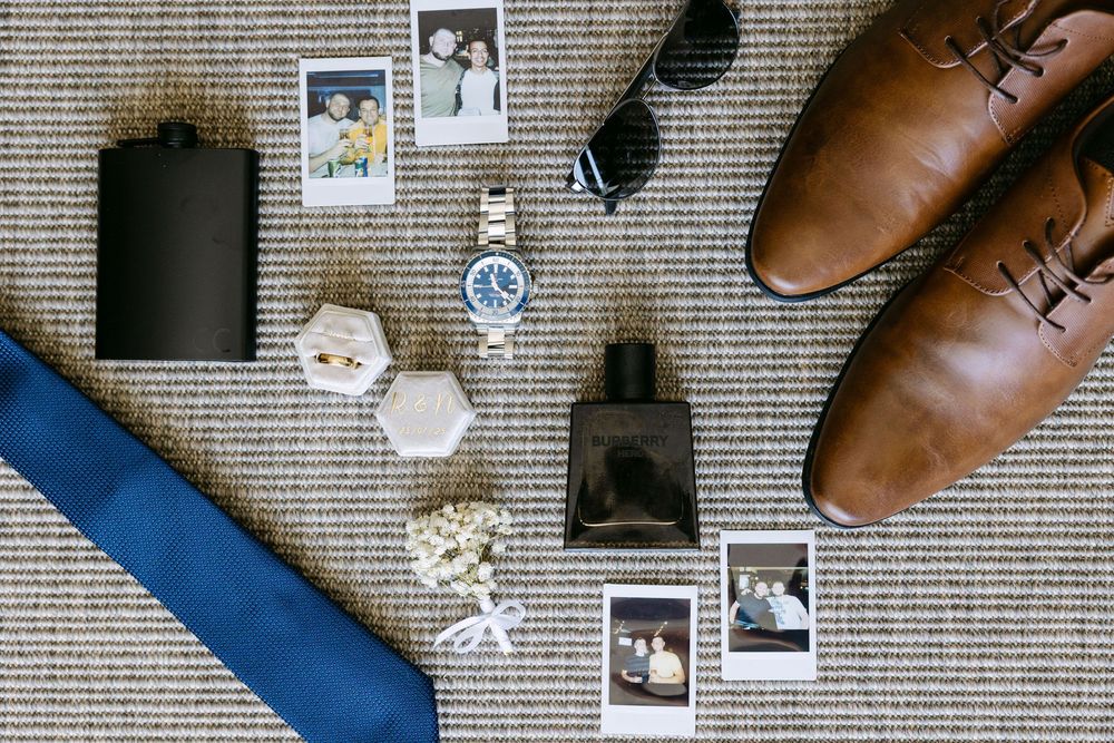 Flatlay of brown leather shoes, sunglasses, and personal accessories arranged on a textured gray background.