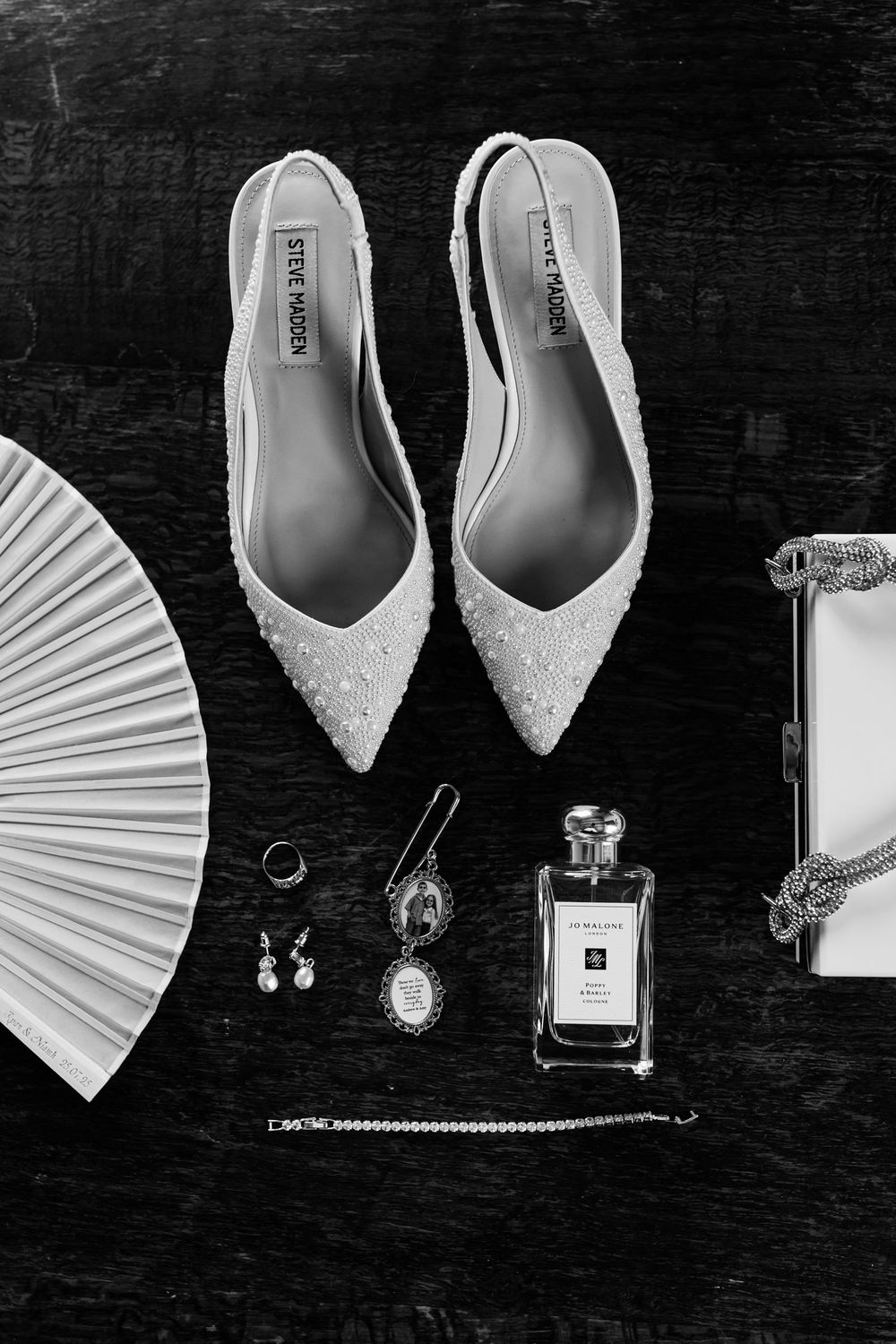 Wedding accessories including white heels, perfume bottle, jewelry, and paper fan arranged on dark surface in black and white.