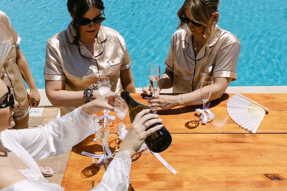 People sitting at a wooden table enjoying drinks and conversation by the pool during a bride preparations for a wedding
