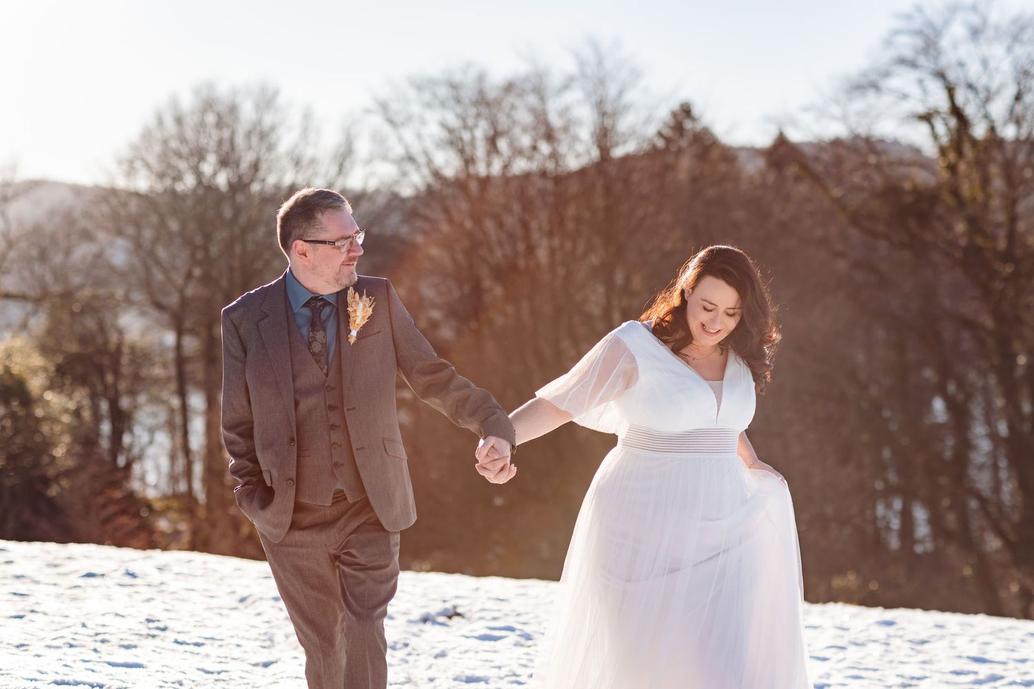 A couple in wedding attire holds hands while walking through a snowy winter landscape at sunset, at Cragwood.
