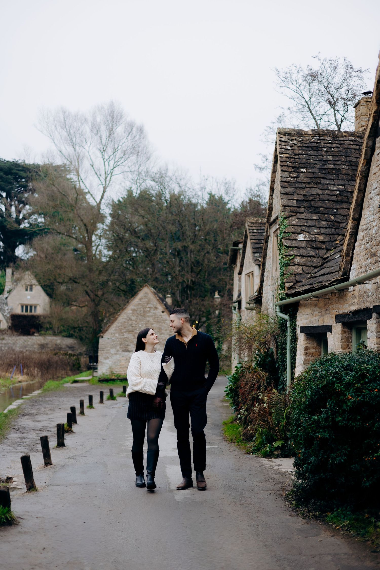 A romantic couple stands together on a wooden footbridge over a stream in a misty forest setting.