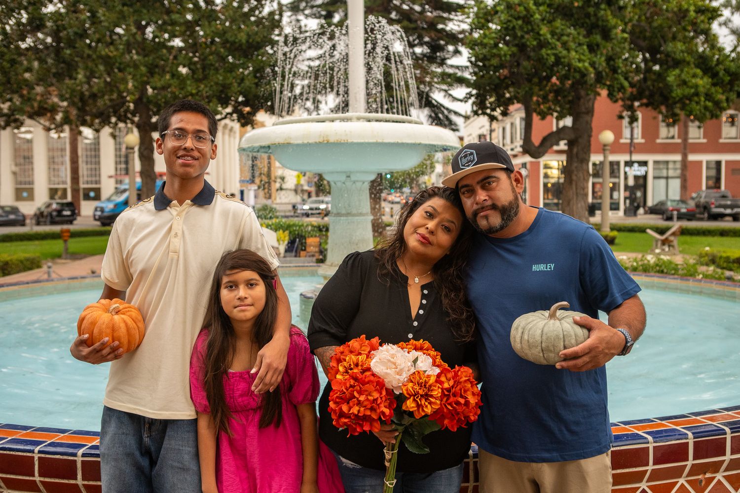 A group stands together by a fountain holding pumpkins and fall flowers on an autumn day.