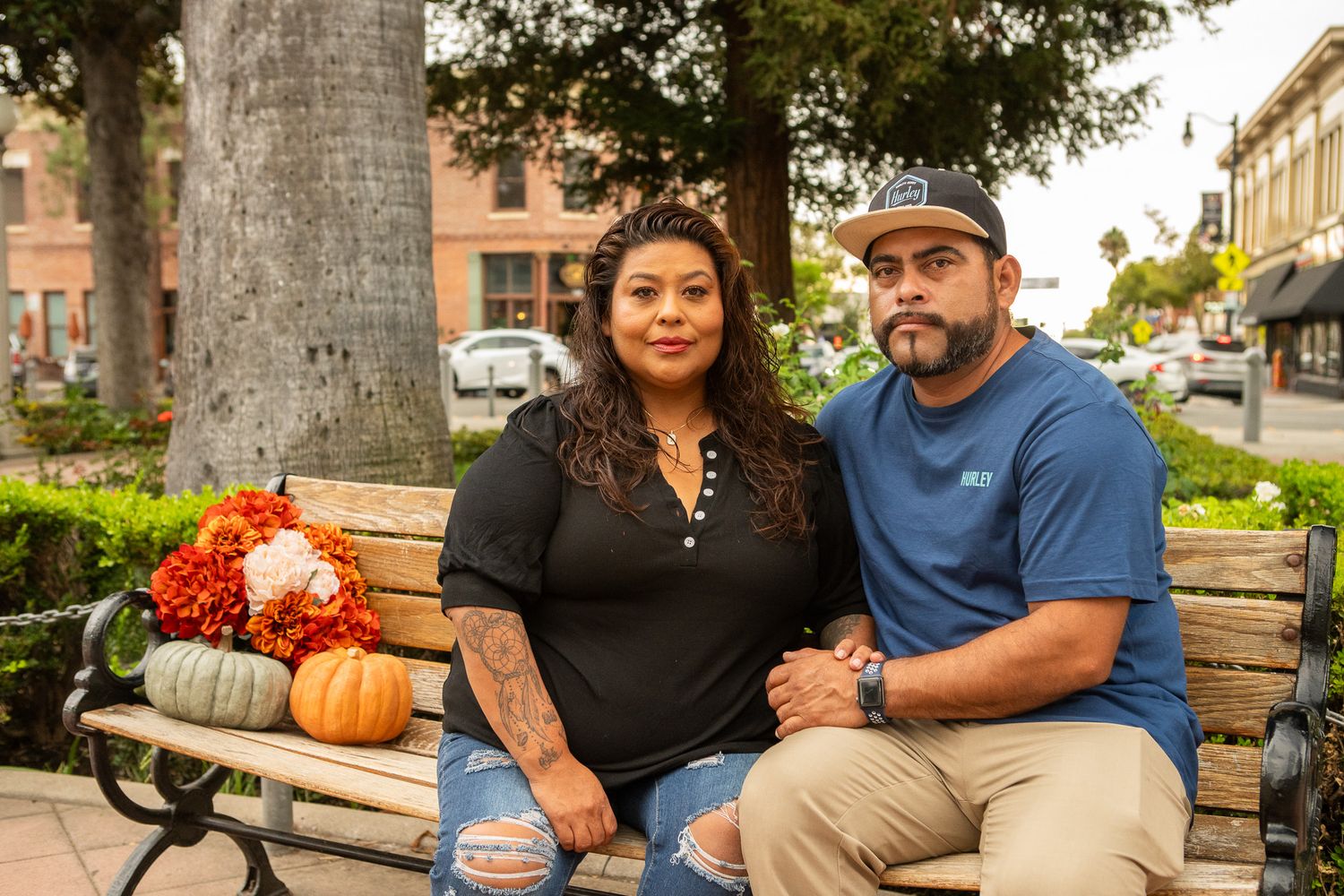 A family sits together on a wooden bench outdoors with autumn pumpkins and decorative arrangements nearby.