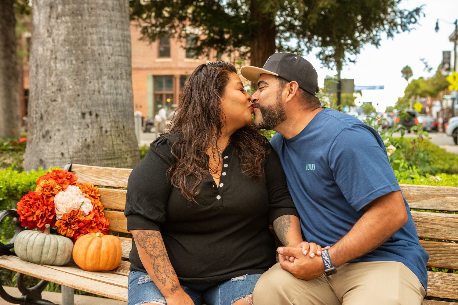 A couple shares a romantic moment on a bench with autumn pumpkins and flowers nearby.