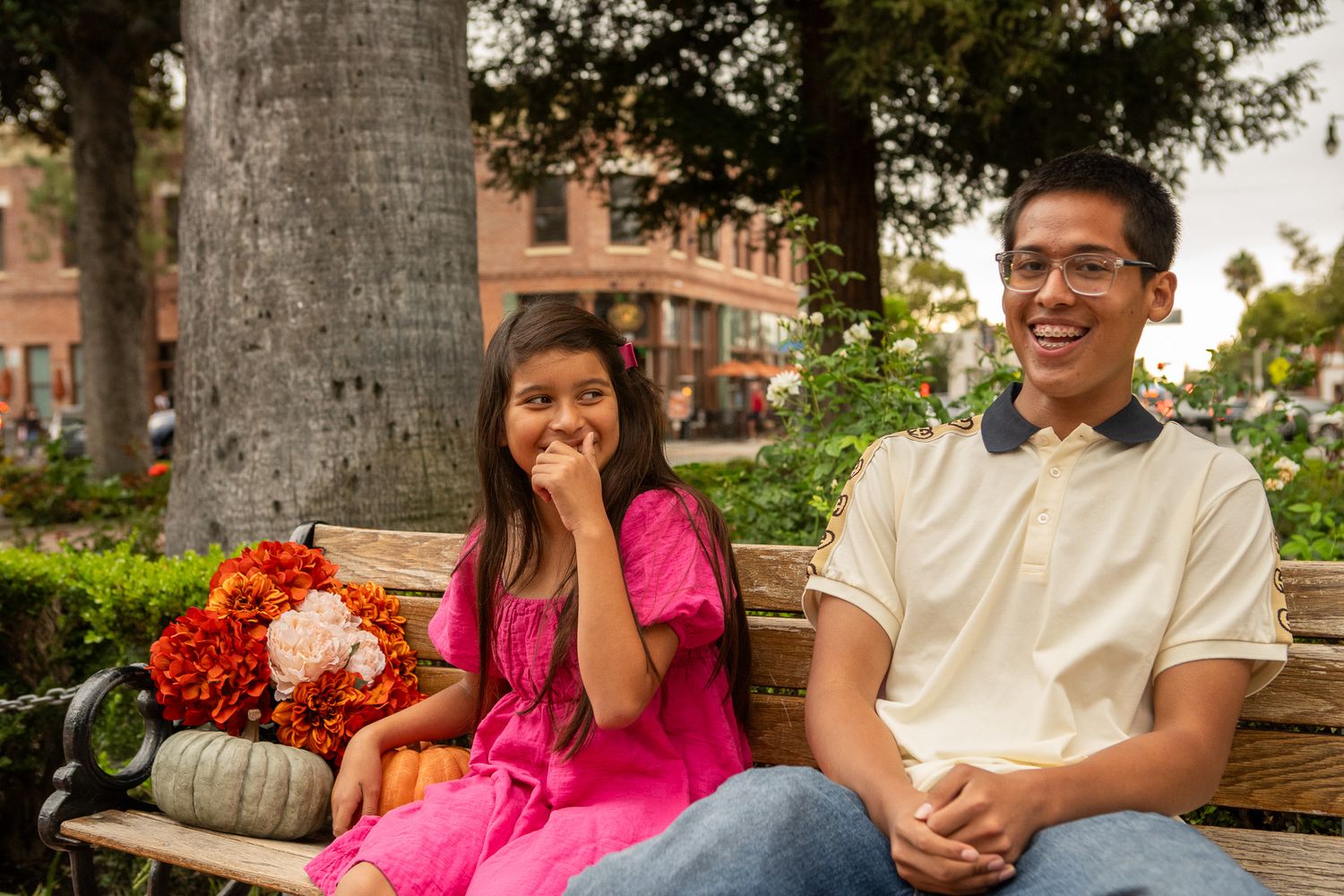 A couple sits on a wooden bench surrounded by palm trees in a casual outdoor setting.