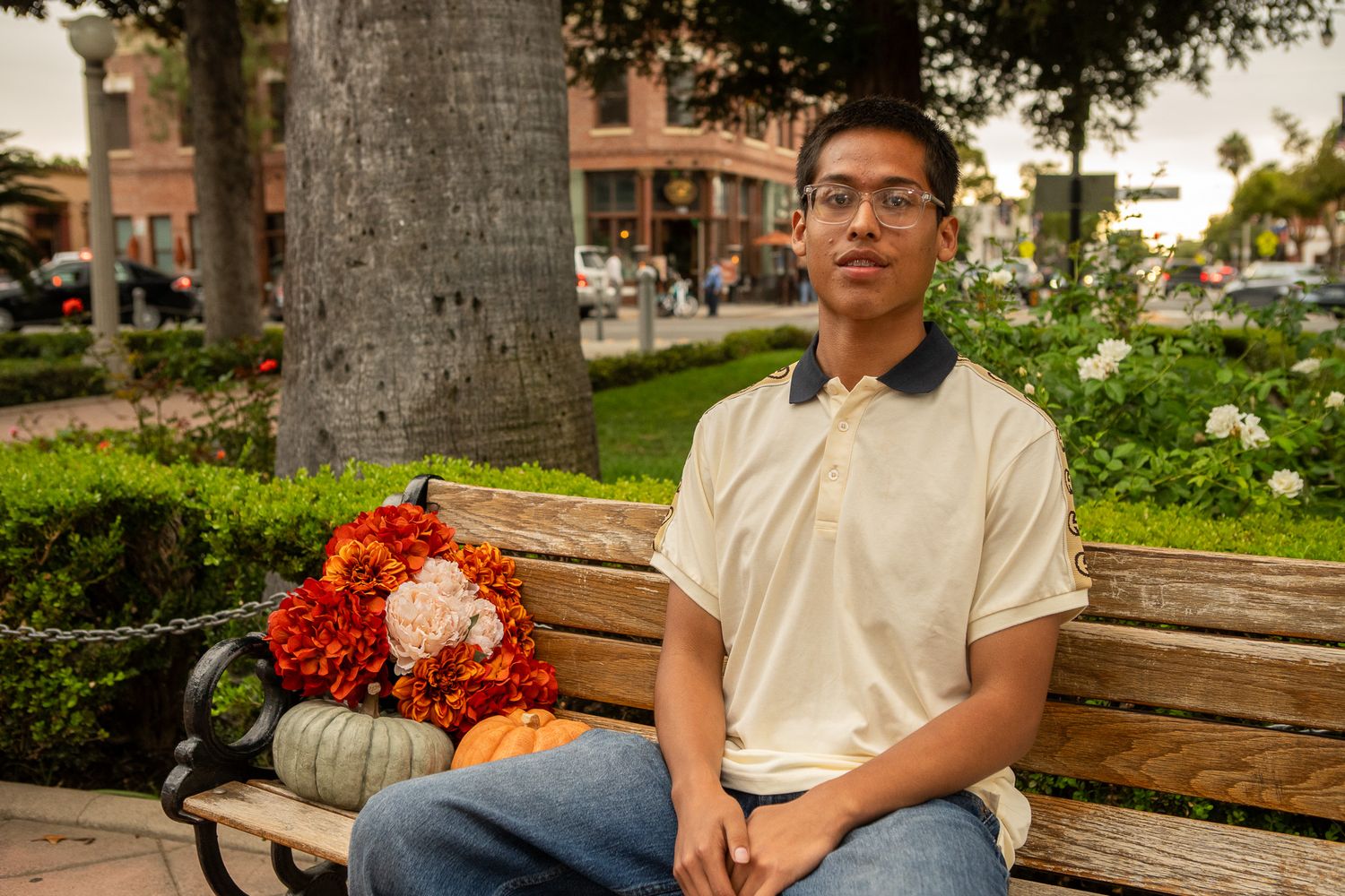 A person sits on a wooden bench near a palm tree with a bouquet of red and orange flowers beside them.