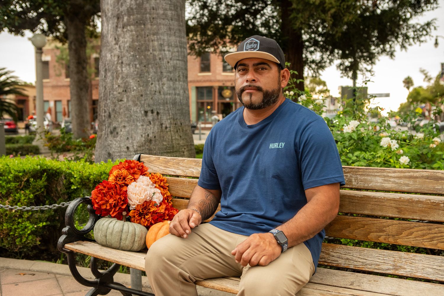 A couple relaxes together on a park bench with lush greenery and buildings in the background.
