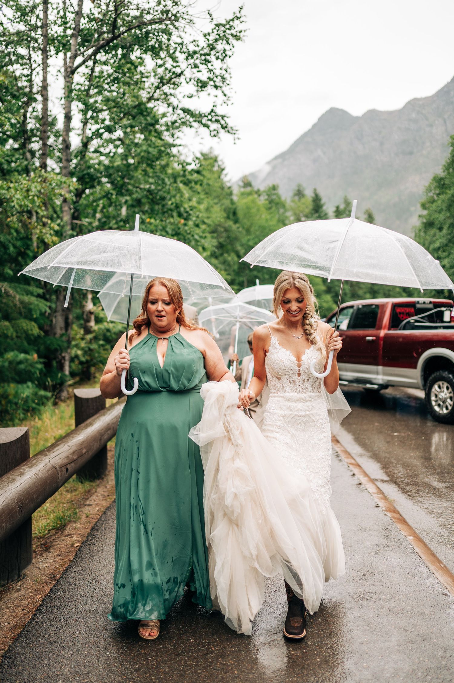 Two people in wedding attire walk together under clear umbrellas on a rainy mountain road.