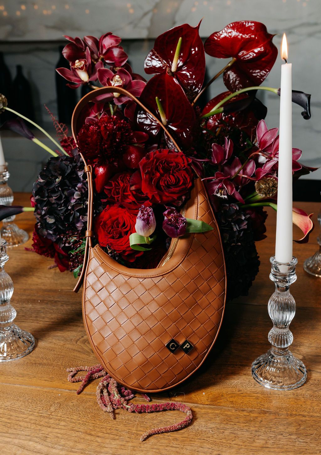 Dark red floral arrangement with roses and orchids in a tan woven leather handbag next to a crystal candlestick on wooden table.