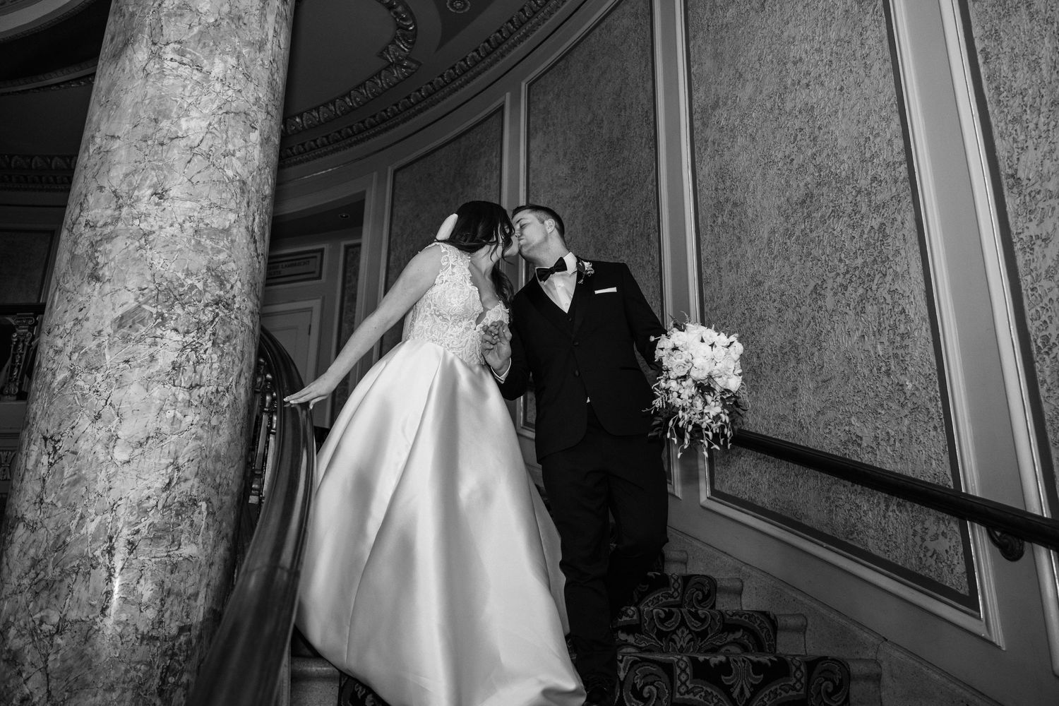 A couple poses in an elegant marble ballroom with ornate chandeliers and classical architecture details.