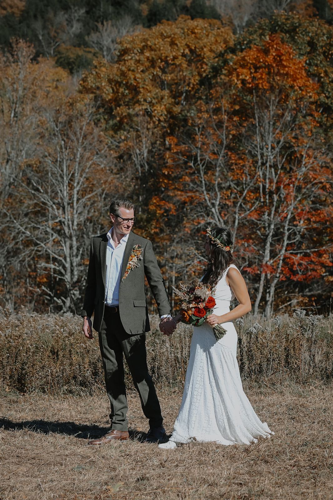 A couple walks together through an autumn field surrounded by trees with vibrant orange and red foliage.