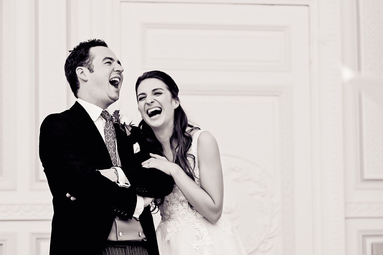A black and white photo captures a joyful moment of laughter between a couple on their wedding day.