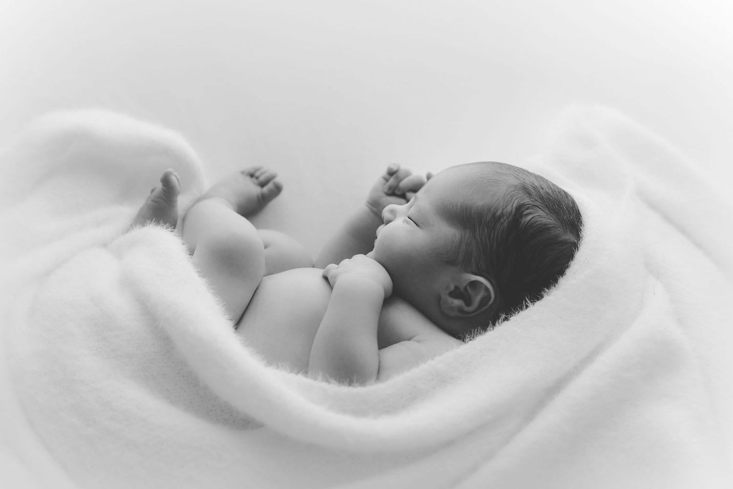 A sleeping newborn baby wrapped in soft white swaddling blankets lies peacefully against a neutral backdrop.