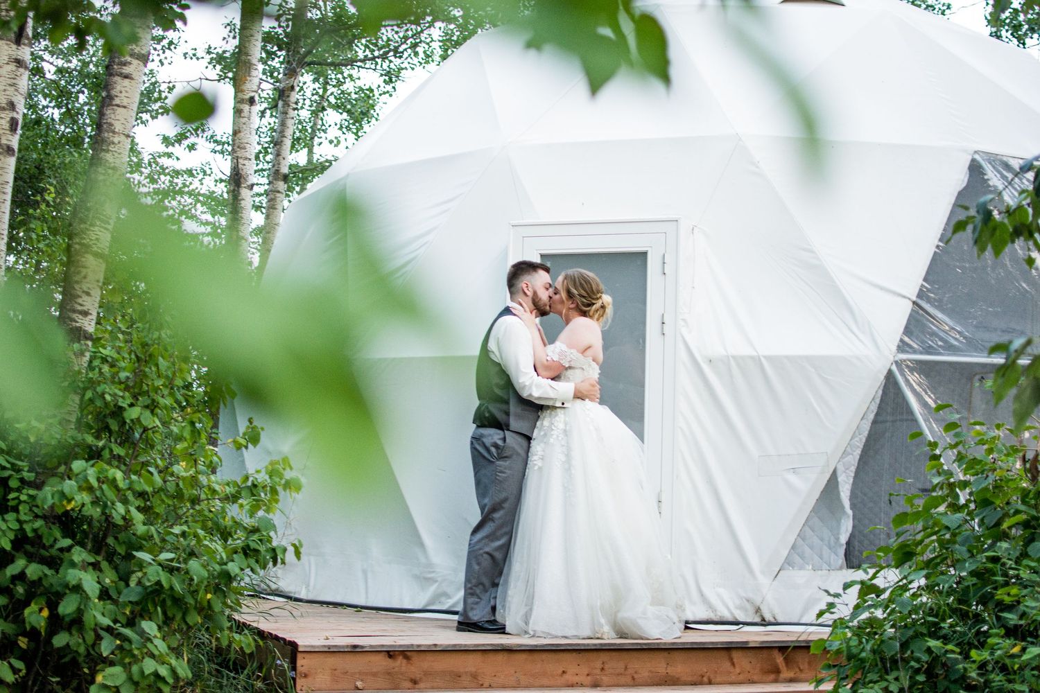 A couple embraces in front of a white geodesic dome structure lush greenery. Garden View Estates, and  Village Vows Seba Beach