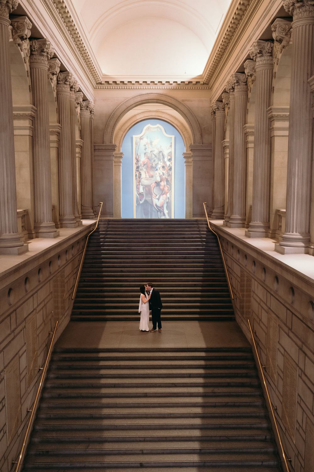 A couple poses on grand marble stairs in a classical architectural setting with arched ceilings.