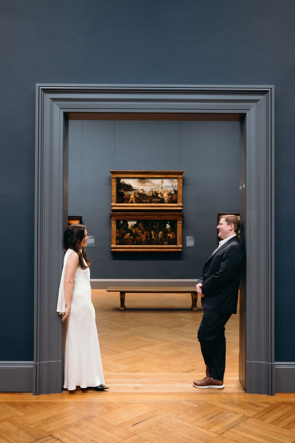 Museum gallery with grey walls displaying classical paintings and herringbone wood floors.