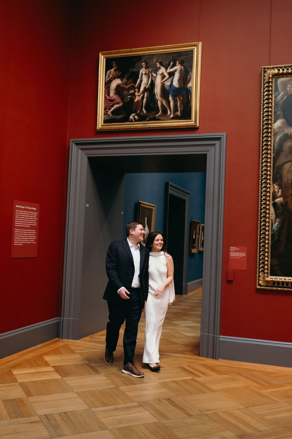 A couple shares a tender moment in an art gallery with red walls and classic paintings.
