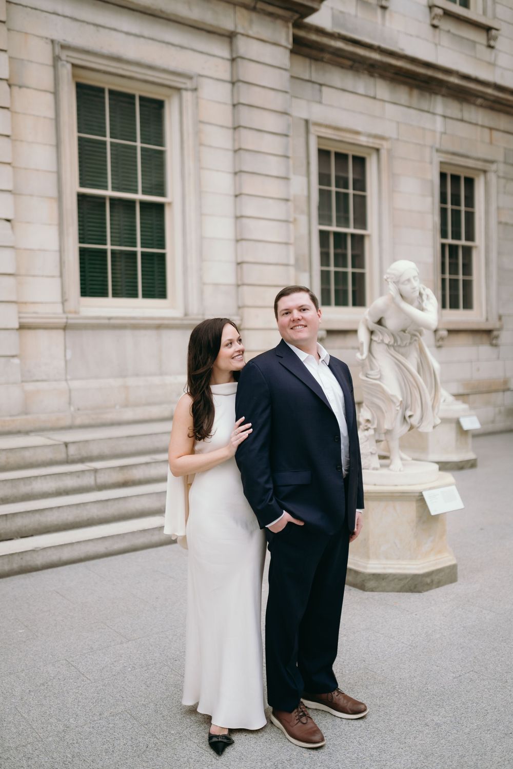 A couple poses playfully outside a classical stone building with decorative sculptures.